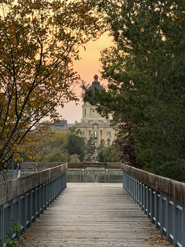 A wooden path leads through autumn trees to the Saskatchewan Legislative Building in Regina, glowing under a colorful sunset sky.