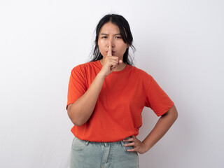 Pretty young Asian woman wearing orange shirt gesturing silence or secret with her finger, isolated by white background.