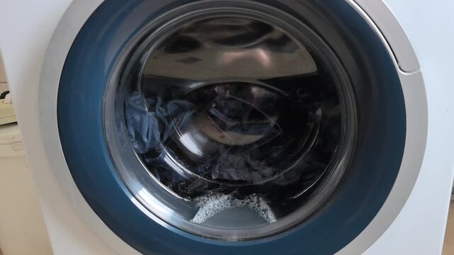 The washing machine is cleaning clothes with soap and water in a home laundry room. Bubbles form in the glass door as the cycle runs. The setting shows a typical weekday afternoon.