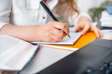 Close up of woman hand writing notes in notebook near open book and laptop, adult student studying online or teacher preparing for distance lesson © 5ph