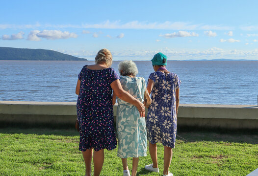 Tr&ecirc;s mulheres caminhando juntas ao longo de um caminho &agrave; beira do lago, vista de tr&aacute;s, representando cuidado, companheirismo e conex&atilde;o entre gera&ccedil;&otilde;es.