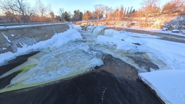 A fast-flowing river with patches of churning white water breaks through thick ice and heavy snow banks in a winter landscape