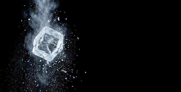 A single, shimmering ice cube shatters into frosty particles and mist against a stark black background, creating a dynamic visual effect.