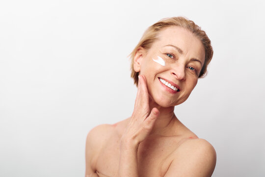 Smiling middle aged woman applying face cream on cheek with hand touching skin. Natural skincare concept with healthy glowing skin and positive self care on white background.