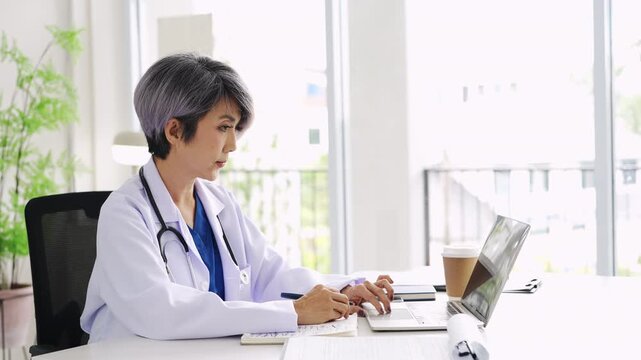 Senior middle aged female medical worker in modern clinic wearing white doctor's gown coat using laptop writing health personal data, consulting remotely. Telemedicine healthcare concept.