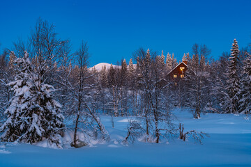 House stands in winter forest of Sweden with snow-covered trees, blue sky, and distant mountains during evening light