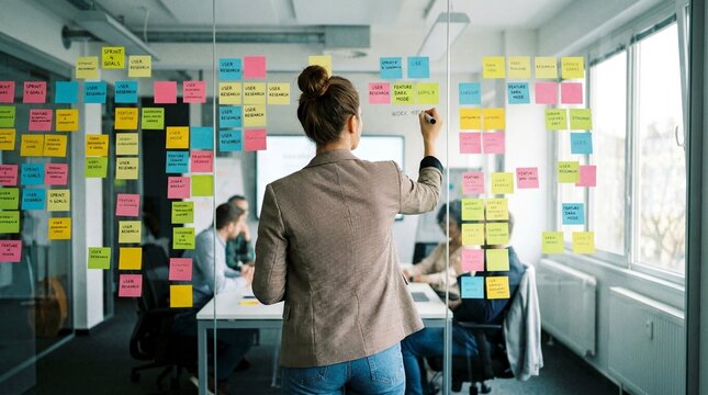 Focused young professional woman stands with her back to the camera while writing new ideas on a clear glass wall covered with colorful sticky notes for agile planning.
