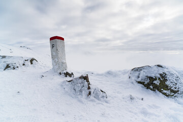 Lonely border post rising from an icy mountain crest, coated with frost and snow, sharply contrasting with white terrain as thick fog and clouds swallow the outlines of distant summits.