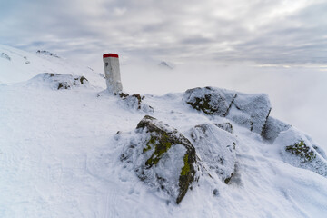 Stone border marker with a red-painted top standing on a snowy mountain ridge, surrounded by frozen rocks, above a deep fog-filled valley and under a layered, overcast winter sky, Tatra National Park