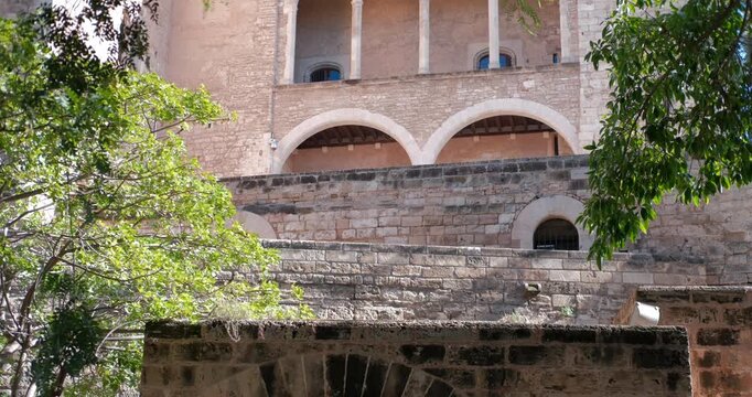 Detailed view of the ancient Almudaina Palace in Palma Majorca Spain. Sunlight brightens the stone facade and green trees.