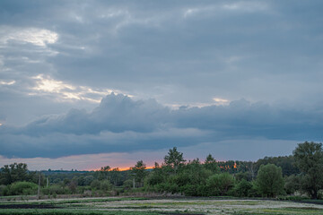 Sunset landscape with storm clouds in blue sky and orange setting sun behind green forest and field of dandelions