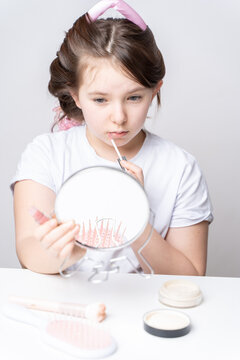Cute little teen girl in curlers paints lips with gloss while looking in mirror at home on white background