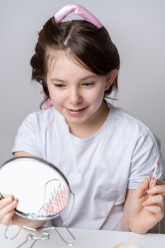 Cute little teen girl in curlers paints lips with gloss while looking in mirror at home on white background