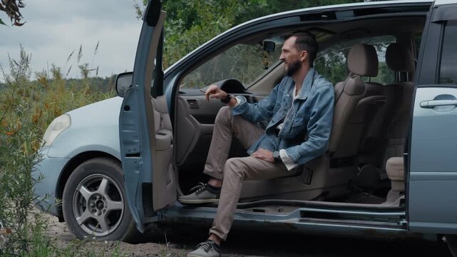 A bearded man in a denim jacket sits alone in his minivan parked in a natural setting during a trip, smoking a cigarette while enjoying a quiet moment.