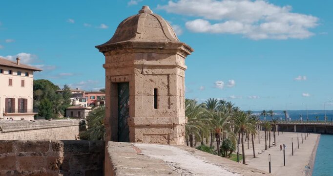 View from Almudaina palace ramparts in Palma, Majorca. A historic guardhouse stands by the sunny harbor promenade and blue sea.
