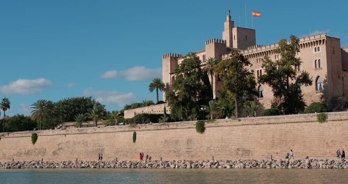 Palma de Mallorca showcases Royal Palace Almudaina and grand Cathedral. People walk along the waterfront on a clear, sunny day in Spain. Pan shot.