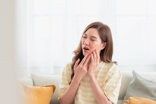 Young Asian woman sitting on a sofa and touching her cheek with an expression of pain and discomfort. Concept of toothache, sensitive teeth, dental problems, and oral healthcare.