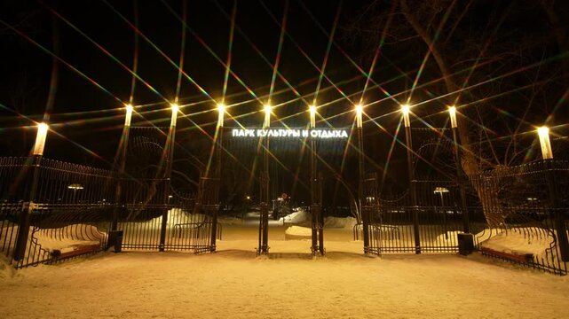 A wide nighttime view of a park entrance featuring a long black metal fence and gate. A glowing sign in Cyrillic letters, hangs above the main opening.