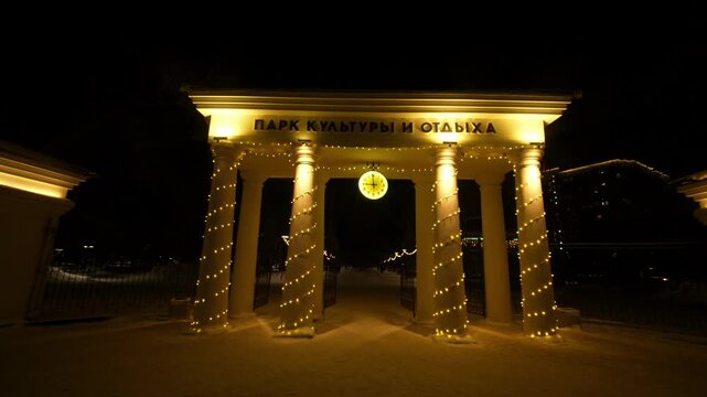 A grand, classical-style entrance gate to a park stands illuminated at night. The structure features four tall columns wrapped in warm golden fairy lights,