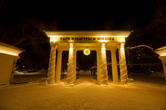Wide angle shot of a grand neoclassical entrance gate to a park at night. The white arch is supported by four columns wrapped in warm golden fairy lights.