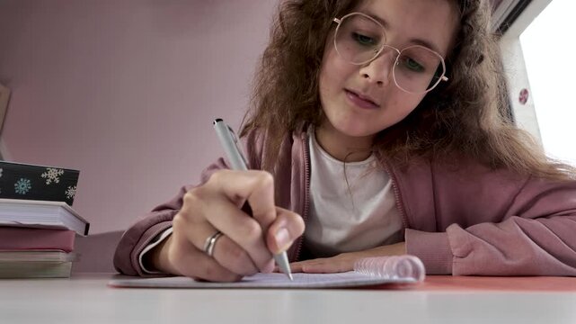 Girl studying diligently, writing in notebook with pen at desk, focused student wearing glasses under soft evening lighting