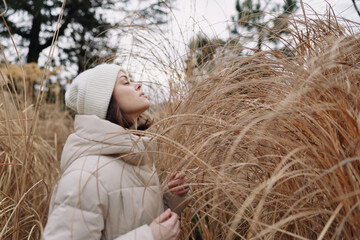 Woman in a puffy coat and knit hat stands among tall dried grass in an open field, capturing a calm winter moment and a intimate connection with nature in a quiet outdoor scene. © SHOTPRIME STUDIO