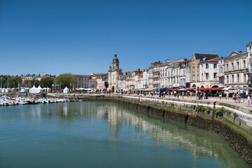 Obraz premium Historic European Port City View - Wide view of a sunny French port with historic waterfront buildings, a prominent clock tower, and boats reflected in the calm water.