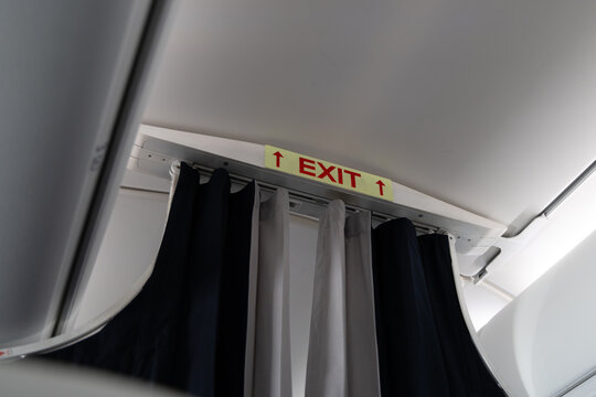 Closeup of an airplane cabin with curtains dividing classes and a red EXIT sign above the aisle, showing passenger safety signage inside a modern commercial aircraft interior