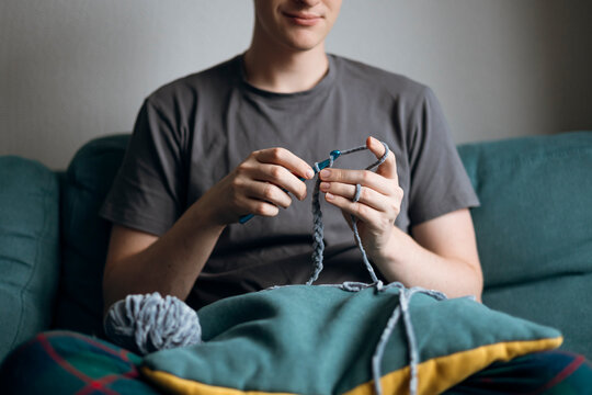 Man crocheting on a couch with teal yarn and pillow