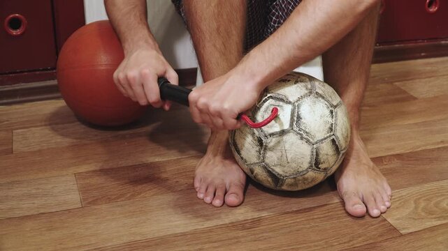 Caucasian man inflates old football with hand pump in room indoor. Process of inflating deflated, torn ball before amateur game at start of season.