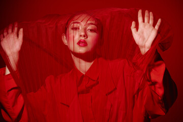 Naklejka na ściany i meble Fashionable young woman in red outfit behind red mesh fabric. Creative beauty concept with strong color contrast on a red background. Artistic high fashion studio shot.