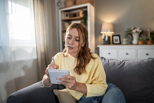 Woman writing notes on notepad at home sofa