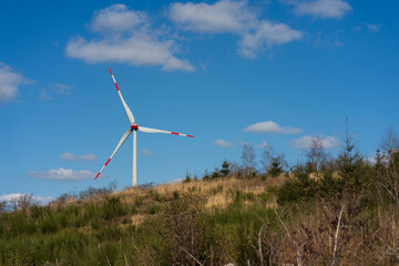 Fototapeta premium A lone wind turbine peeking over a hill against a blue sky with small white clouds, minimalist rural landscape concept showing real life renewable energy in Europe, Germany.