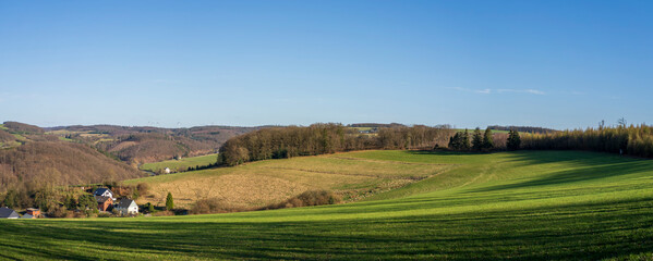 Naklejka premium Panoramic landscape with rolling hills and a distant row of wind turbines in soft haze, wide rural scenery showing real life renewable energy in Europe, Germany