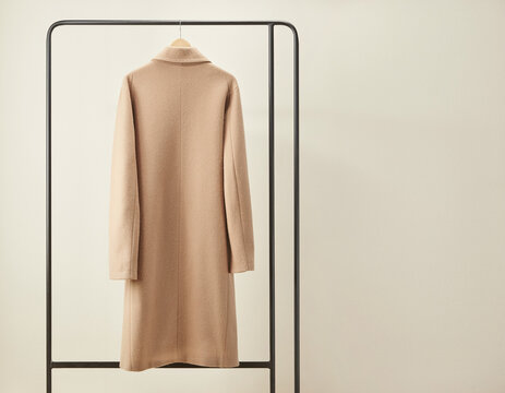 A simple beige woolen jacket suspended from a black metallic hanger. Against a neutral backdrop, the subtle material weave enhances the garment's appeal. 