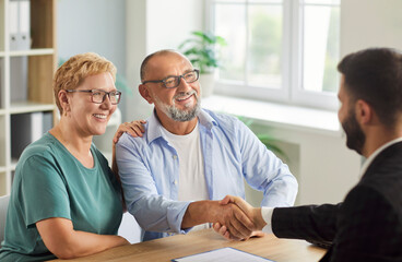 A senior couple meets their advisor in office, shaking hands to finalize a retirement agreement and...