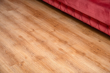 Close-up view of wooden flooring with visible grain texture beside a red upholstered sofa. Ideal for interior design, home decor, real estate, flooring and lifestyle concepts with copy space.