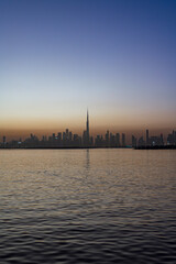 Obraz premium Mesmerizing Dubai skyline at dusk: Burj Khalifa silhouetted against a gradient sunset sky, city lights reflecting on calm waters with a lone boat in the foreground.
