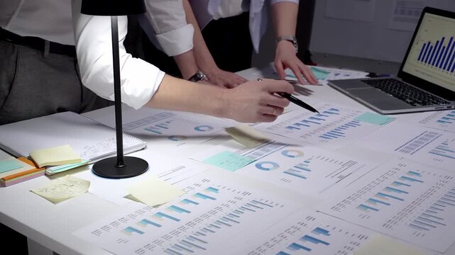 Professional businessman analyzing financial data and charts on laptop and paper documents, writing notes late at night, working overtime in modern office researching company growth and strategy
