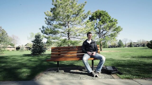 A footage of young man sitting alone on wooden bench in a park on sunny day
