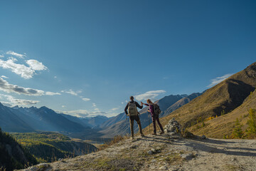 Fototapeta premium Two hikers stand on a mountain pass overlooking a vast valley and snowy peaks under a clear blue sky during autumn travel.