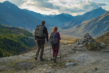 Fototapeta premium Two hikers with backpacks and trekking poles walk along a mountain ridge overlooking a vast autumn valley and snowy peaks under a cloudy sky during an adventure.