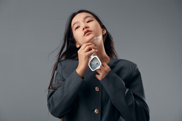 Woman posing with long dark hair and light skin wearing dark jacket holding white sunglasses with hand near chin on gray background, fashion portrait with confident attitude and style.