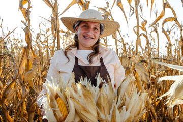 A happy female farmer wearing a straw hat and apron stands in a golden cornfield holding a large...