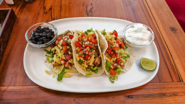 Three fresh tacos served on a white oval platter, topped with vibrant pico de gallo, shredded cheese, and lettuce. Side bowls of black beans and sour cream accompany the meal on a wooden table.