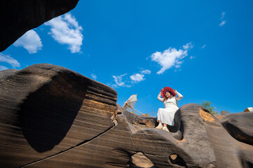 Woman in white dress and red hat sits on large rock formations under bright blue sky with potholes visible in stone Lei Dan Stone Field Thailand