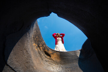 Woman white dress and red jacket looks down into deep hole surrounded by rocky walls clear blue sky with potholes. Lei Dan Stone Field Thailand