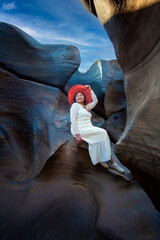 Woman wearing red hat and white dress sits among smooth rock formations blue sky with potholes creating natural shapes Lei Dan Stone Field Thailand