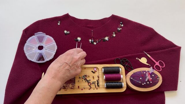 Video of a woman's hand using tweezers to arrange beads and bugle beads on burgundy sweater before sewing them on. Decorating warm women clothing. Beads embroidery as a hobby and pleasure. DIY concept