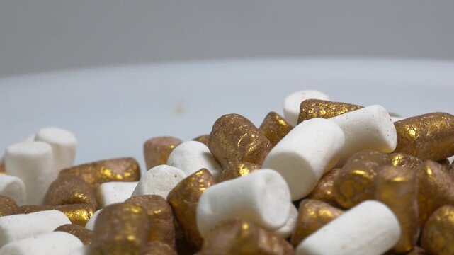 A heap of cylindrical sweet sprinkles in white and gold colors rotates on a plain surface, showcasing their texture and shimmering finish for cake decoration.